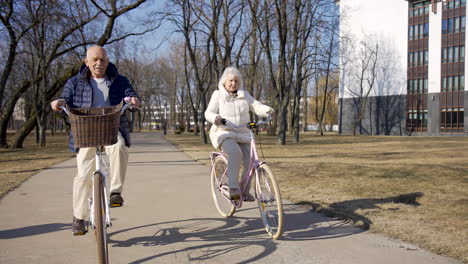 Front-view-of-a-senior-couple-riding-bikes-in-the-park-on-a-winter-day-while-looking-at-camera