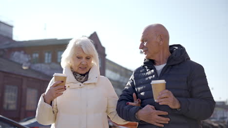 Front-view-of-a-senior-couple-talking-and-walking-down-the-street-on-a-winter-day-while-holding-coffees-to-go