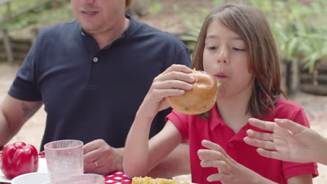 Close-up-of-happy-boy-eating-burger-during-a-picnic-in-the-forest-with-his-family