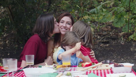 Medium-shot-of-sons-sitting-around-mother,-kissing-her-and-then-looking-at-the-camera-during-a-family-picnic