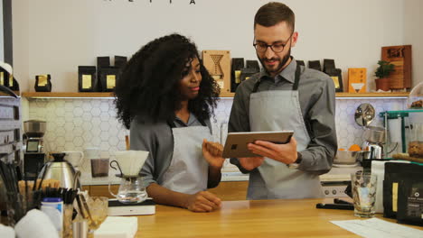 Young-caucasian-barista-showing-to-her-african-american-coworker-the-new-menu-of-cafe-on-a-tablet-during-workday