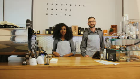 Young-caucasian-barista-and-her-african-american-coworker-posing-for-the-camera-and-crossing-arms-behind-the-bar-of-a-coffee-shop