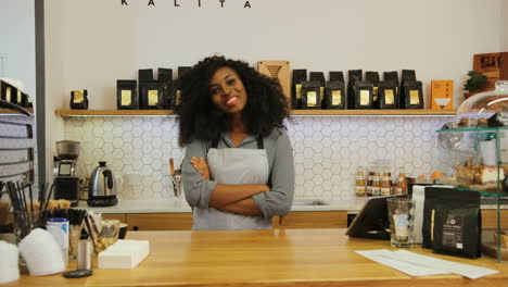 Young-african-american-female-barista-posing-for-the-camera-and-crossing-arms-behind-the-bar-of-a-coffee-shop