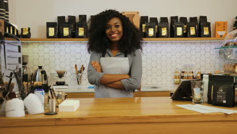 Young-african-american-female-barista-posing-for-the-camera-and-crossing-arms-behind-the-bar-of-a-coffee-shop