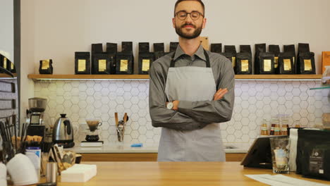 Young-caucasian-male-barista-wearing-glasses-posing-for-the-camera-and-crossing-arms-behind-the-bar-of-a-coffee-shop