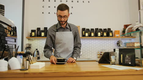 Close-up-view-of-male-barista-in-coffee-shop-making-a-cup-of-coffee-for-the-visitor-and-looking-at-the-camera