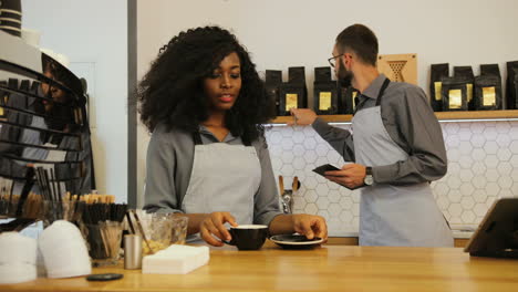 African-american-young-female-barista-making-coffee-behind-the-bar-while-her-coworker-is-checking-the-assortment-of-coffee-and-tea-in-coffee-shop