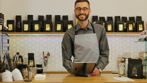 Caucasian-male-barista-with-beard-and-glasses-using-the-tablet,-then-he-looks-at-the-camera-and-smiles-in-a-coffee-shop
