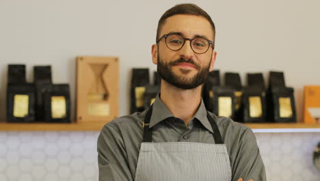 Close-up-view-of-happy-male-barista-wearing-glasses-at-the-coffee-shop-possing-and-smiling-at-the-camera
