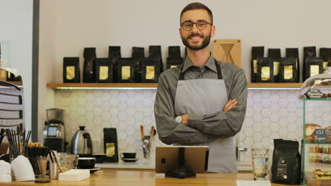 Caucasian-male-barista-with-beard-and-glasses-using-the-tablet,-then-he-looks-at-the-camera-and-smiles-with-crossed-arms-in-cofee-shop
