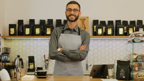 Close-up-view-of-happy-male-barista-wearing-glases-at-the-coffee-shop-possing-and-smiling-at-the-camera-with-crossed-arms