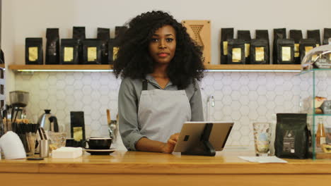 African-american-female-barista-with-curly-hair-using-the-tablet,-then-he-looks-at-the-camera-and-smiles