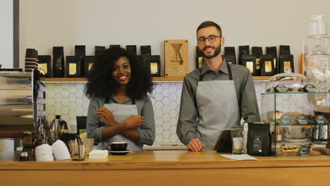 African-american-female-barista-and-her-male-coworker-smiling-and-looking-at-the-camera-with-crossed-arms-behind-the-bar-of-coffee-shop