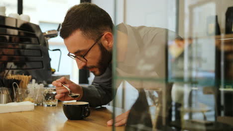 Close-up-view-of-caucasian-male-barista-with-beard-and-glasses-tasting-the-fresh-coffee-in-a-coffee-shop
