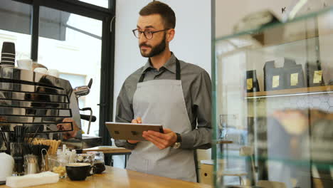 Caucasian-male-barista-with-beard-and-glasses-using-a-tablet-in-a-coffee-shop
