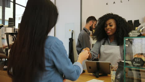 Rear-view-of-young-caucasian-woman-paying-for-a-coffe-with-credit-card-while-african-american-female-barista-serving-her-the-coffee-and-her-male-coworker-is-making-coffee