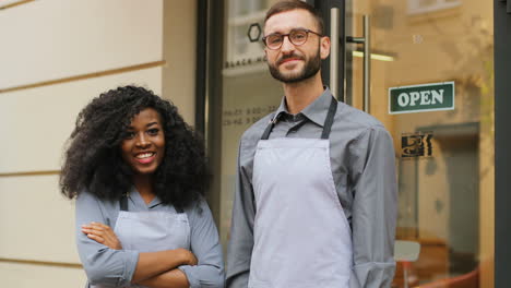 Caucasian-male-barista-and-african-american-female-barista-standing-on-the-door-of-a-modern-cafe,-smiling-and-looking-at-the-camera