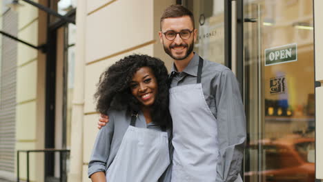 Portrait-of-female-and-male-waiters-hugging-and-smiling-at-camera-outside-of-a-modern-coffee-shop