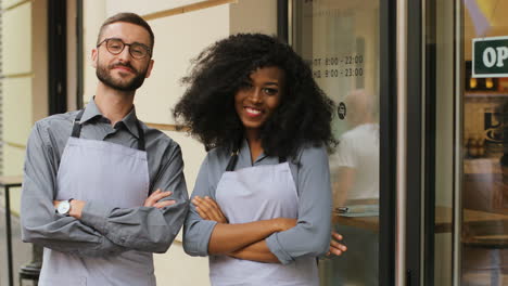Caucasian-male-barista-and-african-american-female-barista-standing-on-the-door-of-a-modern-cafe,-smiling-and-looking-at-the-camera