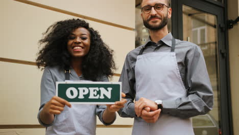 Portrait-of-smiling-caucasian-waiter-and-african-american-waitress-standing-holding-signboard-open"-outside-of-coffee-shop"