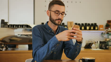 Young-caucasian-man-in-glasses-using-smartphone-while-sitting-at-the-table-in-a-coffee-shop