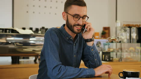 Young-caucasian-man-in-glasses-talking-on-smartphone-while-sitting-at-the-table-with-cup-of-coffee-in-a-coffee-shop
