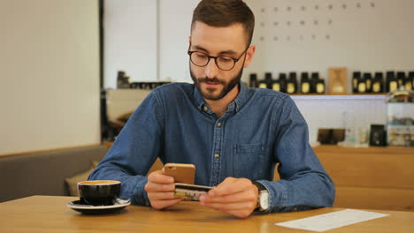 Hipster-man-in-glasses-using-credit-card-for-online-shopping-with-smartphone-while-sitting-a-in-a-cafe