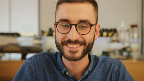 Portrait-of-young-man-with-beard-and-glasses-looking-at-the-camera-and-smiling-in-a-cafe