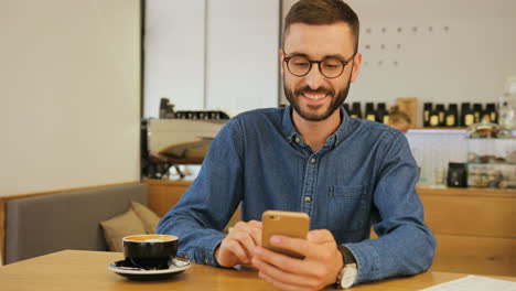 Young-caucasian-man-in-glasses-using-smartphone-for-watching-a-funny-video-while-sitting-at-a-table-in-a-cafe