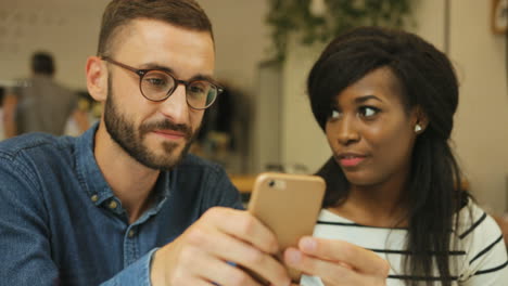 Close-up-view-of-young-caucasian-man-using-smartphone-and-showing-a-video-to-african-american-woman-a-in-a-coffee-shop