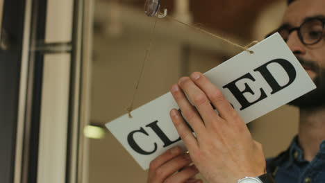 Close-up-view-of-young-man-turning-over-a-closed"-signboard-in-coffee-shop-door"