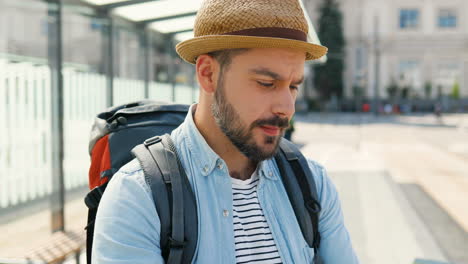 Young-handsome-Caucasian-traveller-wearing-hat-with-backpack-holding-city-map-and-planning-route-in-the-street
