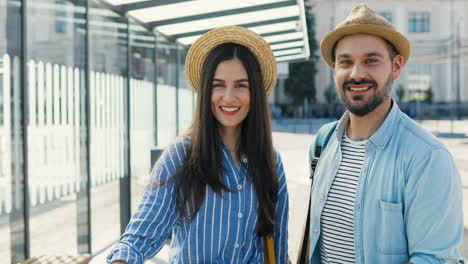 Portrait-of-Caucasian-young-happy-couple-of-travellers-in-hats-with-suitcases-standing-at-bus-stop-and-smiling-to-camera-cheerfully