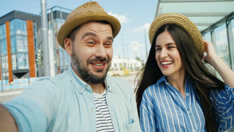 Close-up-view-of-Caucasian-young-happy-couple-of-tourists-in-hats-talking,-waving-hands-and-smiling-at-the-camera-outdoors-in-summer