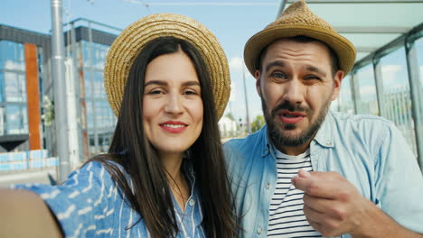 Close-up-view-of-Caucasian-young-happy-couple-of-tourists-in-hats-talking,-waving-hands-and-smiling-at-the-camera-outdoors-in-summer