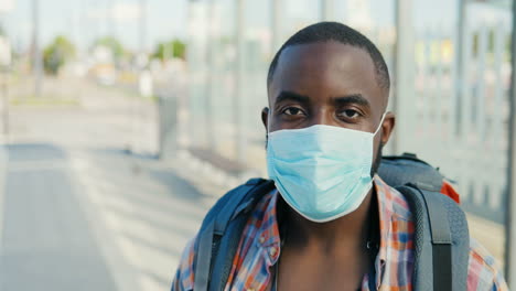 Portrait-of-young-African-American-traveller-man-with-backpack-and-wearing-medical-mask-looking-at-camera-outdoors