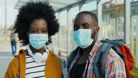 Portrait-of-young-African-American-travellers-couple-with-backpack-and-wearing-medical-mask-looking-at-camera-outdoors