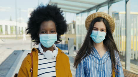 Portrait-of-young-African-American-and-caucasian-women-travellers-with-backpack-and-wearing-medical-mask-looking-at-camera-outdoors