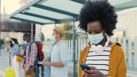 African-american-woman-in-facial-mask-and-headphones-using-smartphone-at-bus-stop-while-others-travellers-waiting-for-transport
