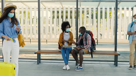 African-american-young-happy-man-and-woman-travellers-in-facial-masks-sitting-at-bus-stop-talking-and-watching-a-map-and-a-tablet-to-plan-a-route