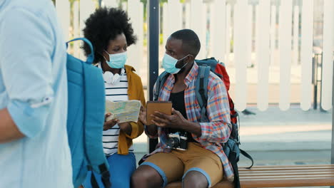 African-american-young-happy-man-and-woman-travellers-in-facial-masks-sitting-at-bus-stop-talking-and-watching-a-map-and-a-tablet-to-plan-a-route