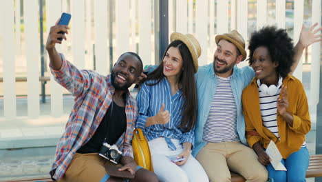 Grupo-Multiétnico-De-Viajeros-Sentados-En-La-Parada-Del-Autobús,-Sonriendo-Y-Haciéndose-Un-Selfie