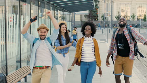 Group-of-multiethnic-friends-travellers-having-fun,-running-and-jumping-at-bus-station