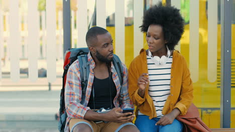African-american-young-happy-man-and-woman-travellers-sitting-at-bus-stop-talking-to-plan-a-route