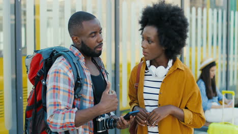 African-american-young-happy-man-and-woman-travellers-at-bus-stop-talking-and-using-smartphone-to-plan-a-route