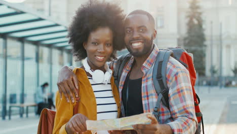 Jóvenes-Afroamericanos-Felices-Hombres-Y-Mujeres-Viajeros-En-La-Parada-De-Autobús-Viendo-Un-Mapa-Y-Mirando-A-La-Cámara