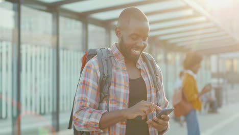 Junger-Gutaussehender-Afroamerikanischer-Mann-Mit-Rucksack-SMS-Nachricht-Auf-Smartphone-Und-Fröhlichem-Lächeln-Am-Bahnhof