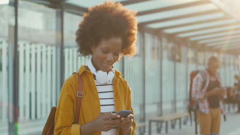 Young-African-American-woman-traveller-with-backpack-texting-message-on-smartphone-and-smiling-cheerfully-at-train-station