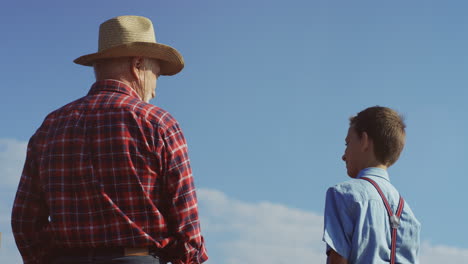 Rear-view-of-a-grandfather-in-a-hat-and-cute-teen-grandson-talking-outdoors-with-blue-sky-on-the-background