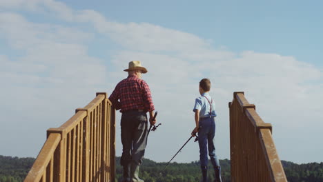 Rear-view-of-senior-man-in-a-hat-standing-on-the-wooden-bridge-with-little-boy-and-fishing-with-rods-in-hands
